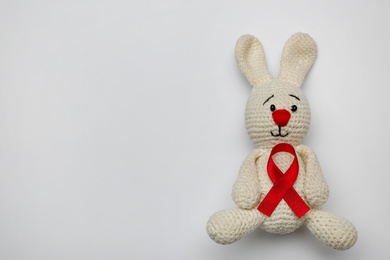 Photo of Cute knitted toy bunny with red ribbon on light grey background, top view and space for text. AIDS disease awareness