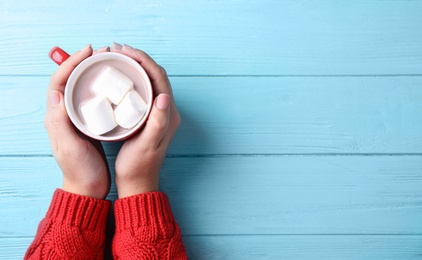 Woman holding cup of tasty cocoa with marshmallows on blue wooden table, top view. Space for text