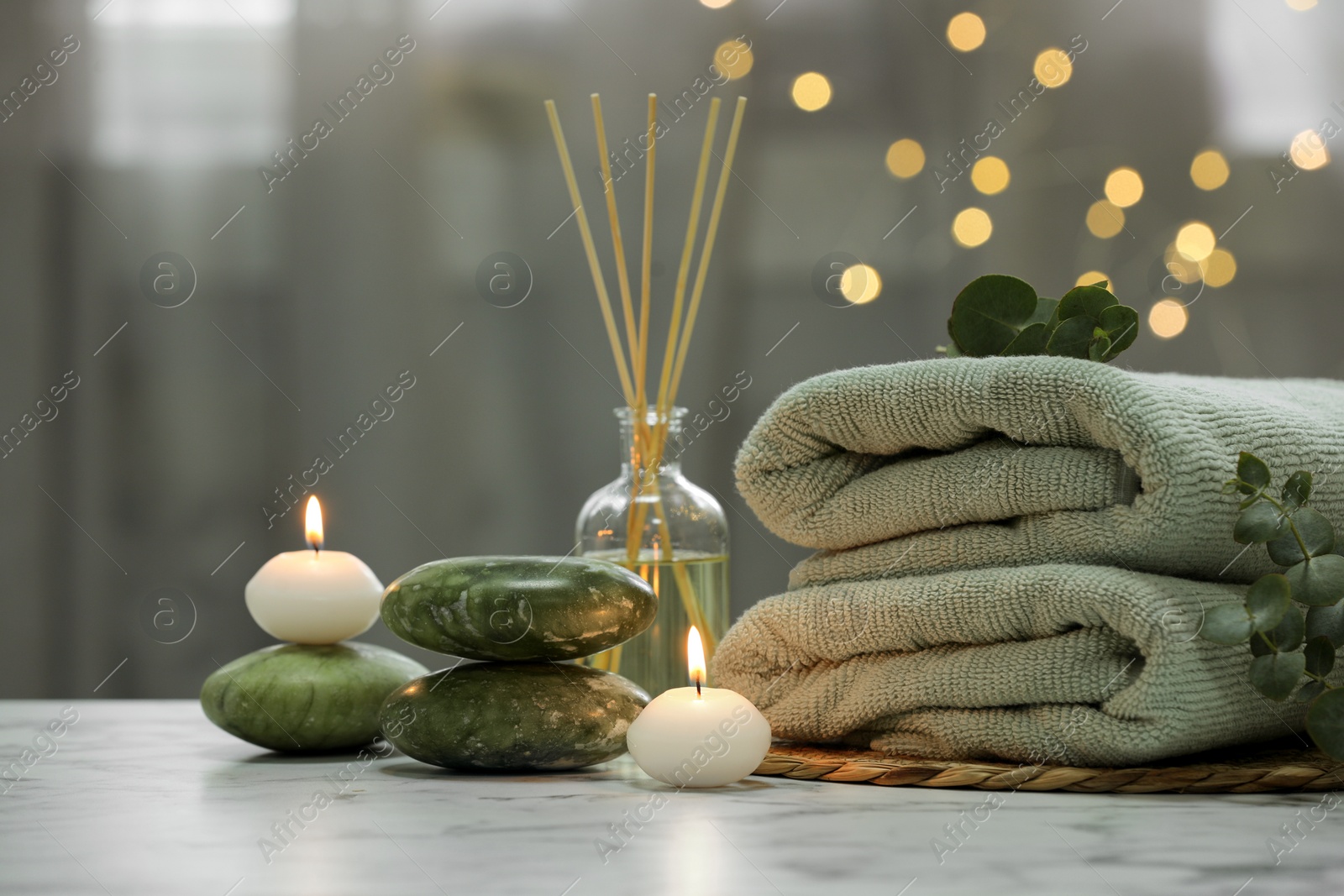 Photo of Spa composition. Towels, stones, reed air freshener and burning candles on white marble table against blurred lights