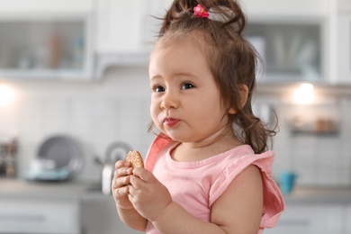 Adorable baby girl eating tasty cookie on blurred background