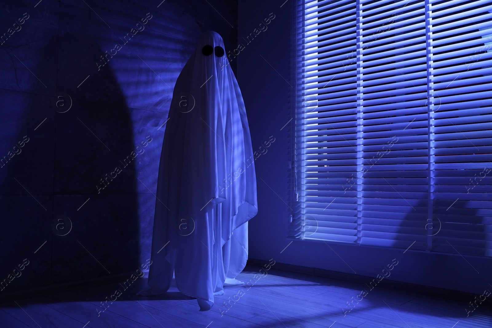 Photo of Creepy ghost. Woman covered with sheet near window in blue light