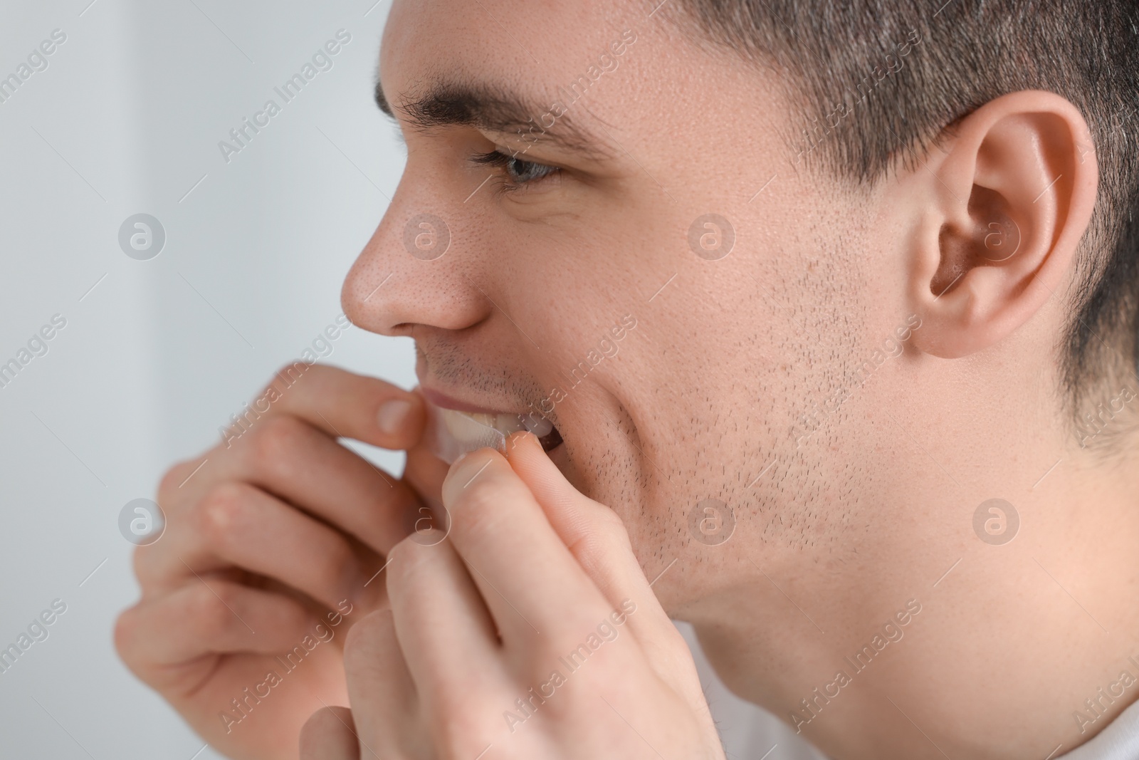 Photo of Young man applying whitening strip on his teeth indoors, closeup