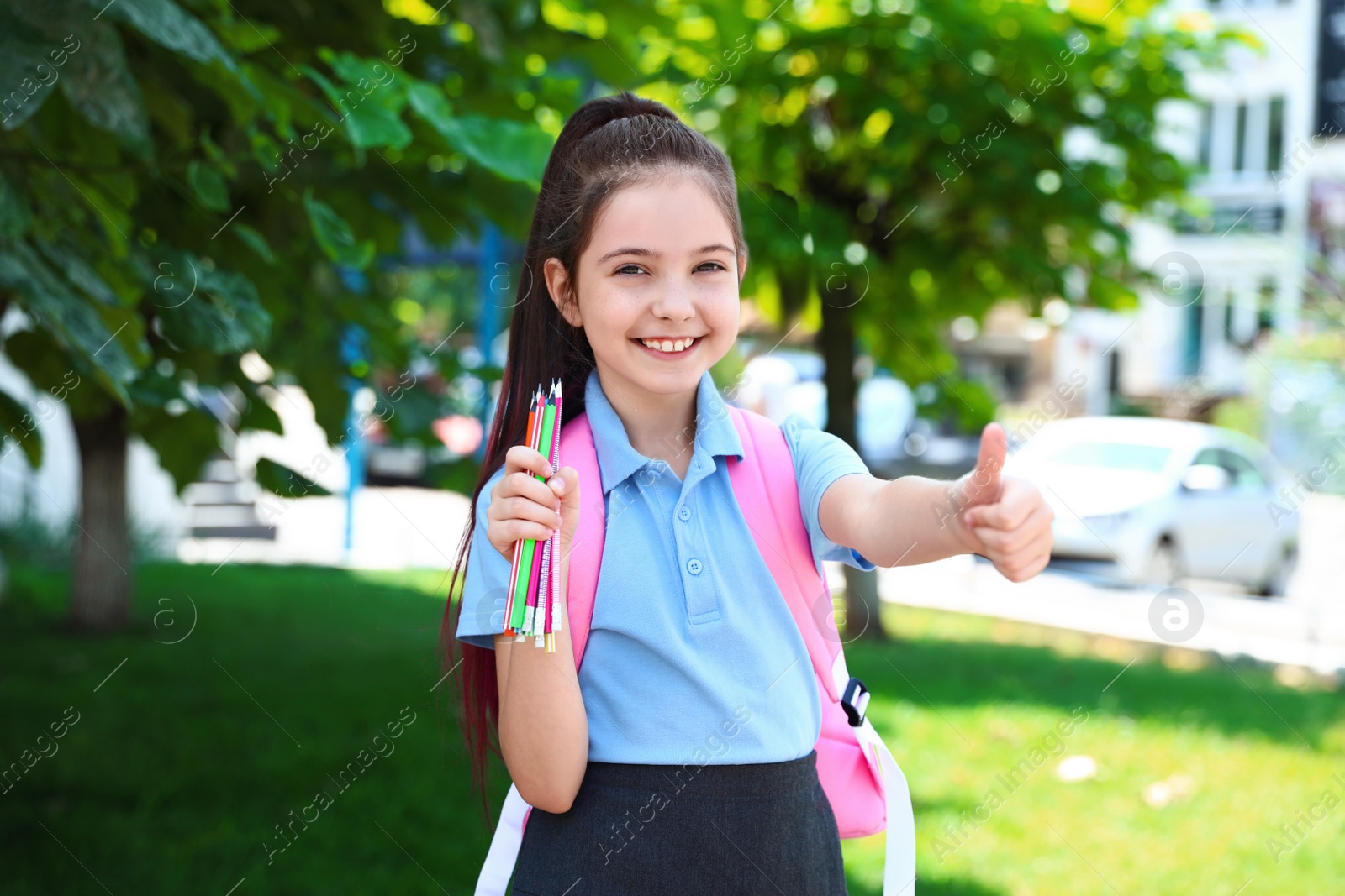 Photo of Cute little girl in school uniform with backpack and stationery showing thumbs-up on street