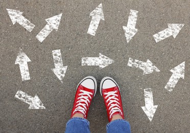 Image of Choosing future profession. Girl standing in front of drawn signs on asphalt, top view. Arrows pointing in different directions symbolizing diversity of opportunities