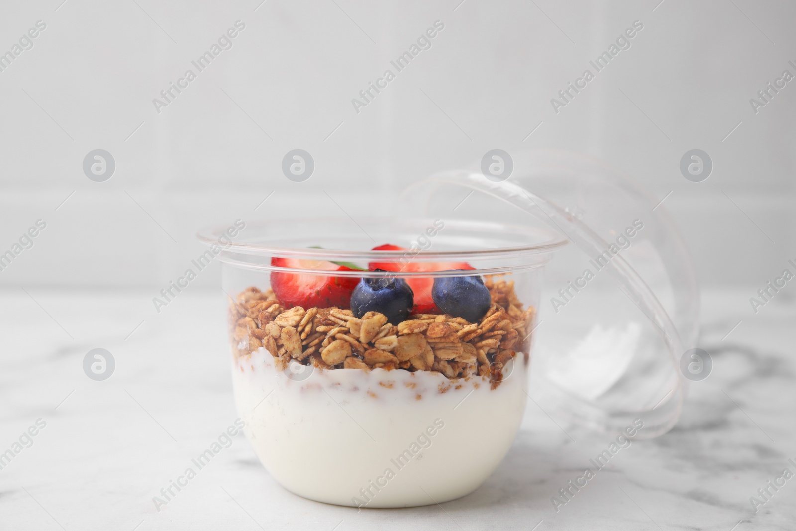 Photo of Tasty granola with berries and yogurt in plastic cup on white marble table