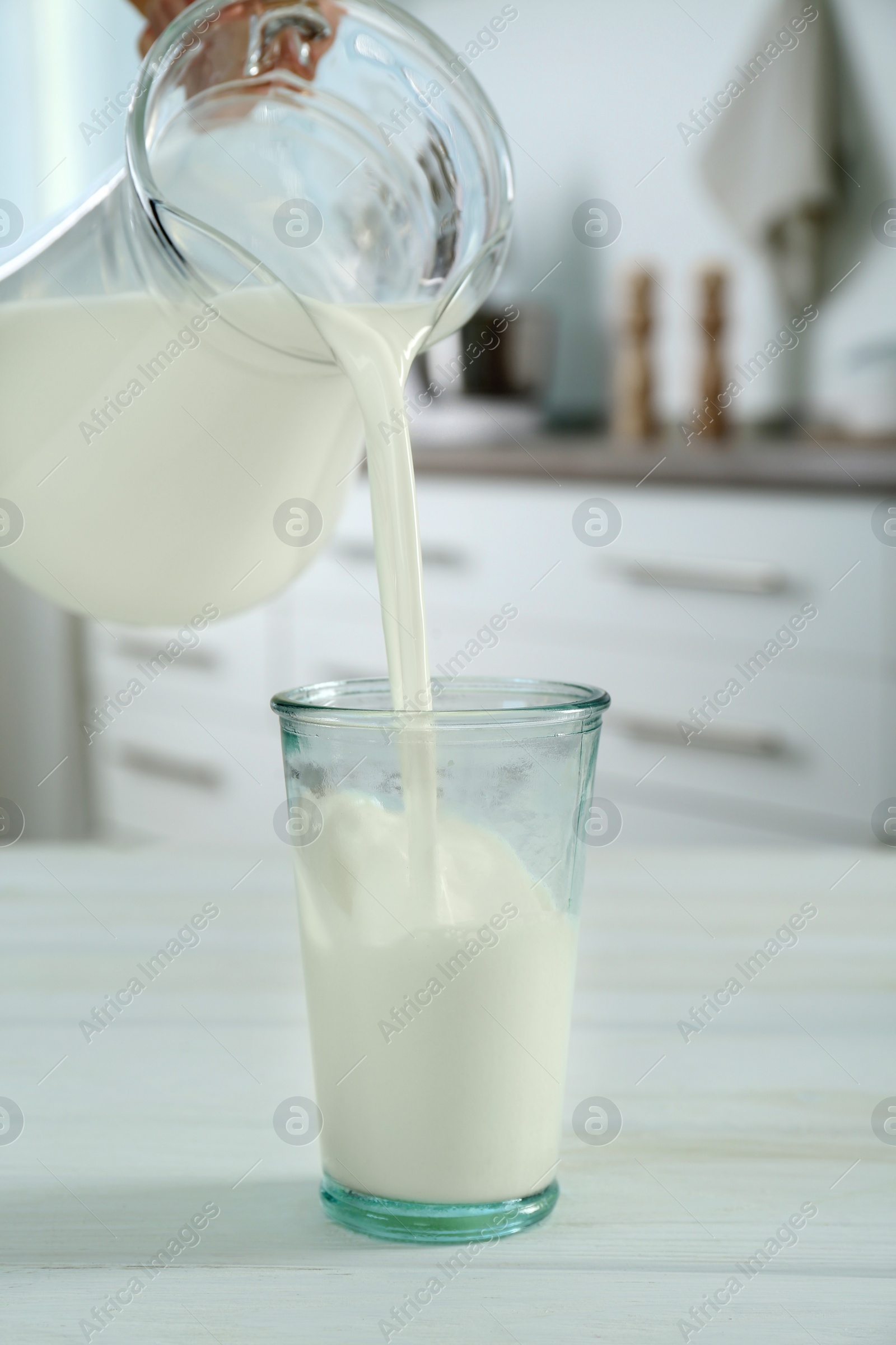 Photo of Woman pouring milk from jug into glass at white wooden table in kitchen, closeup