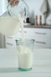 Woman pouring milk from jug into glass at white wooden table in kitchen, closeup