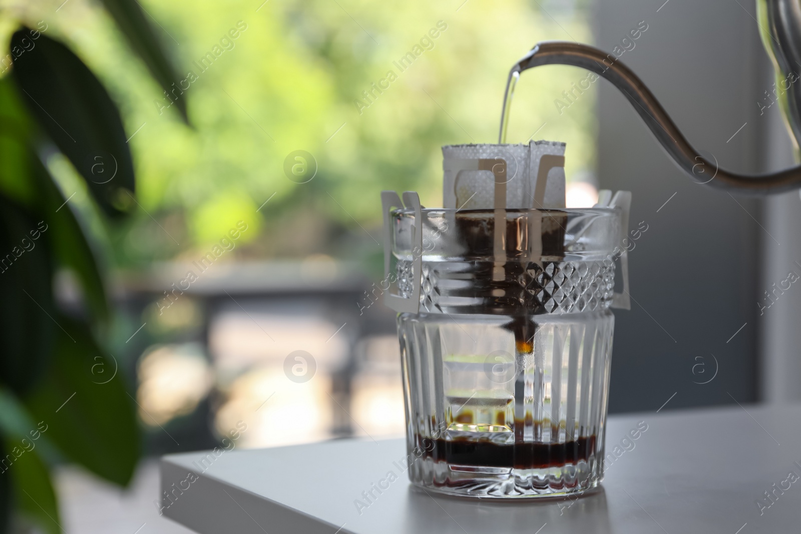 Photo of Pouring hot water into glass with drip coffee bag from kettle on light grey table, closeup. Space for text