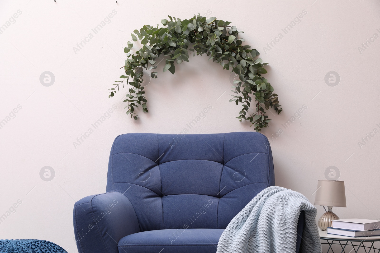 Photo of Beautiful garland made of eucalyptus branches hanging above armchair on white wall indoors