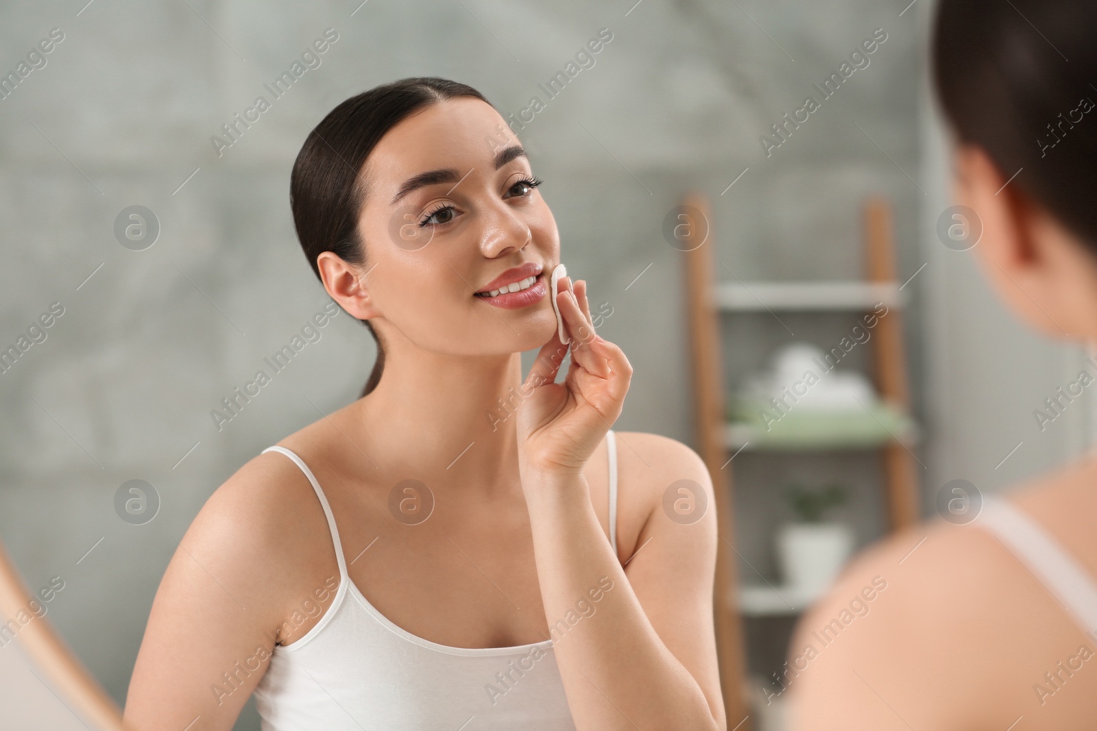 Photo of Beautiful woman removing makeup with cotton pad near mirror indoors