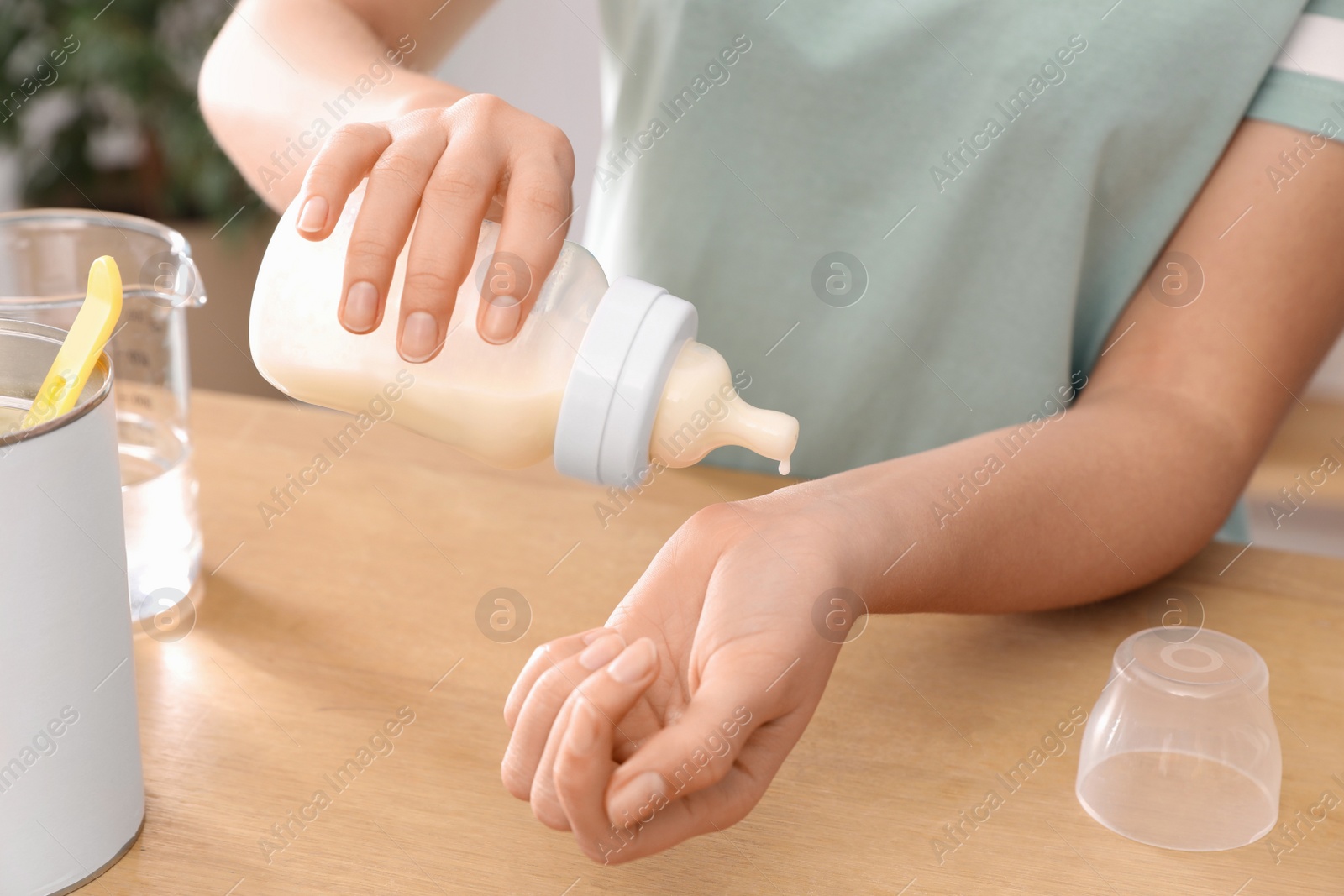 Photo of Woman checking temperature of infant formula at table indoors, closeup. Baby milk