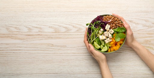 Image of Woman holding delicious vegan bowl with tofu cheese, microgreens and buckwheat at white wooden table, top view. Banner design with space for text