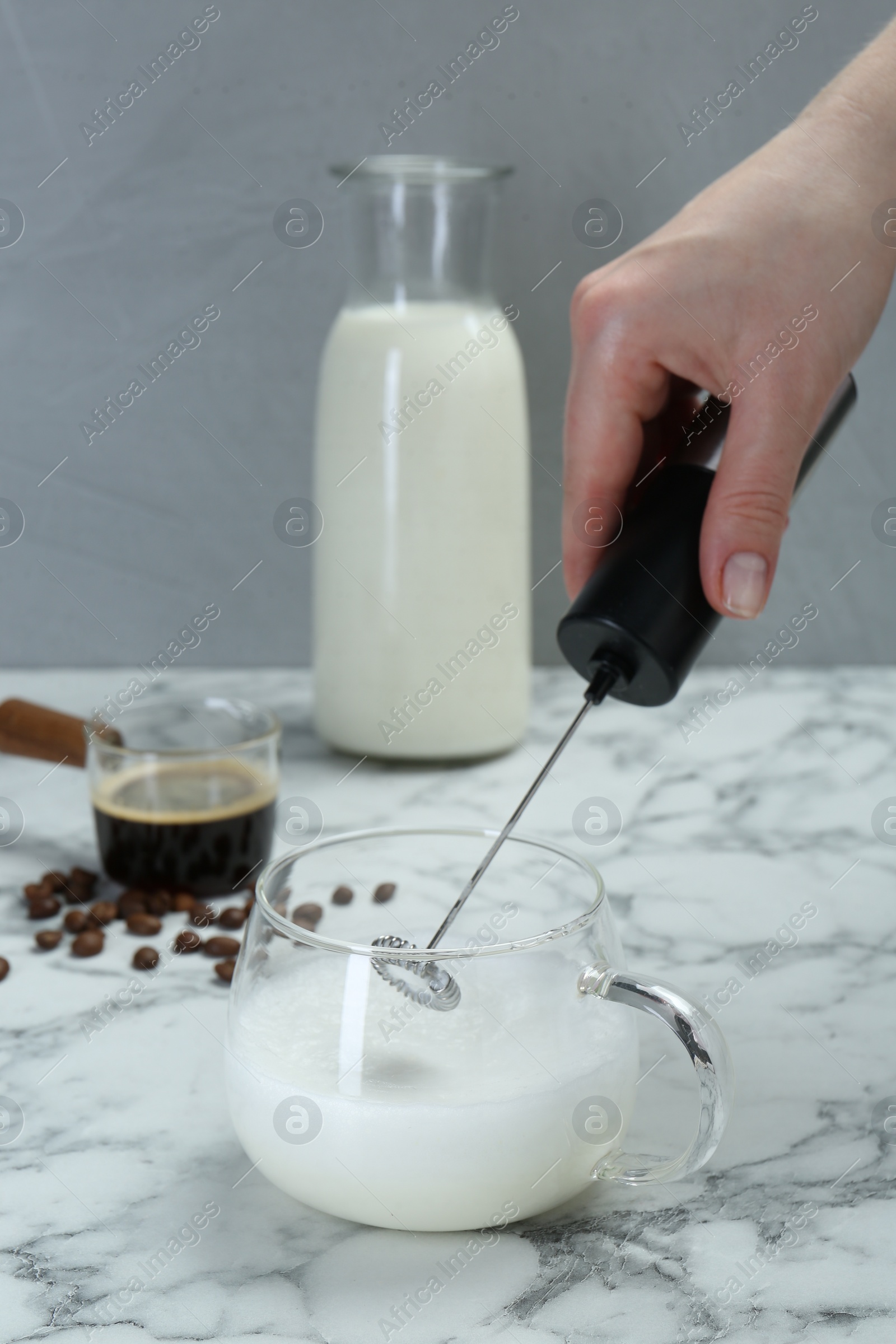 Photo of Woman whisking milk in cup with mini mixer (milk frother) at white marble table, closeup