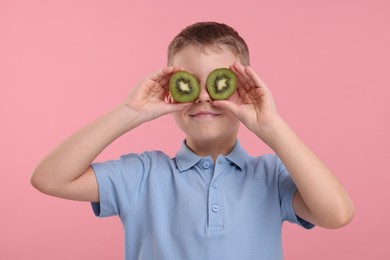 Photo of Boy covering eyes with halves of fresh kiwi on pink background