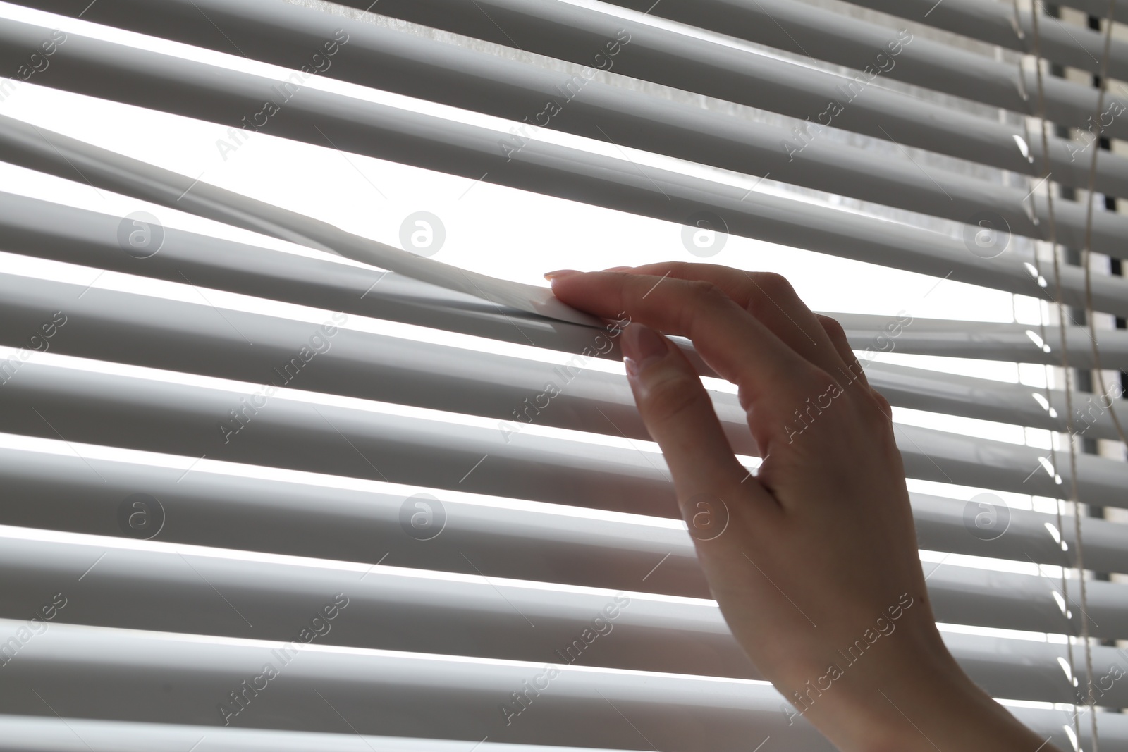 Photo of Woman separating slats of white blinds indoors, closeup