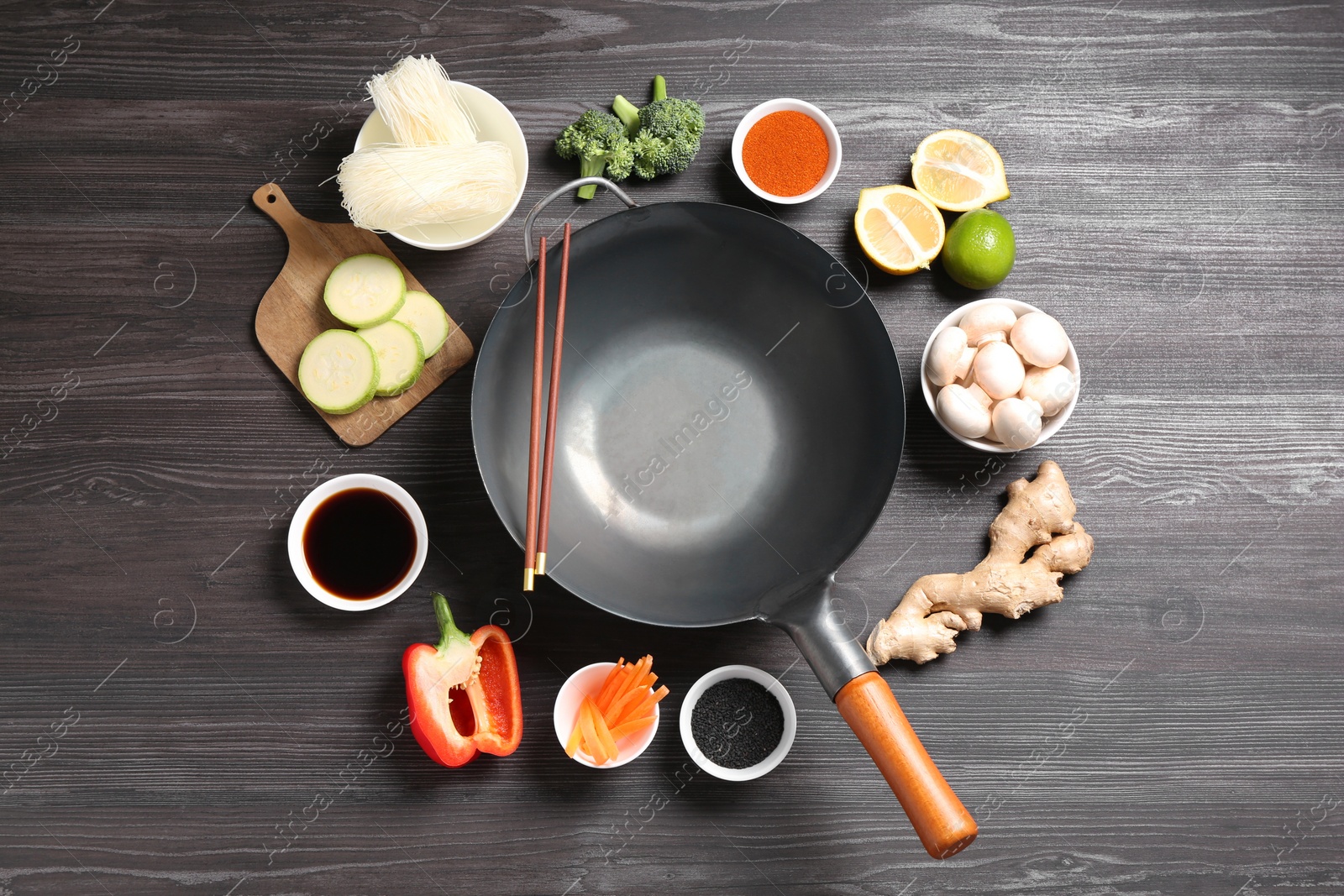 Photo of Empty iron wok and chopsticks surrounded by ingredients on dark grey wooden table, flat lay