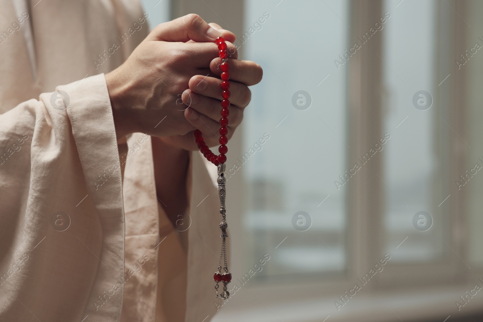 Photo of Muslim man with misbaha praying near window indoors, closeup. Space for text