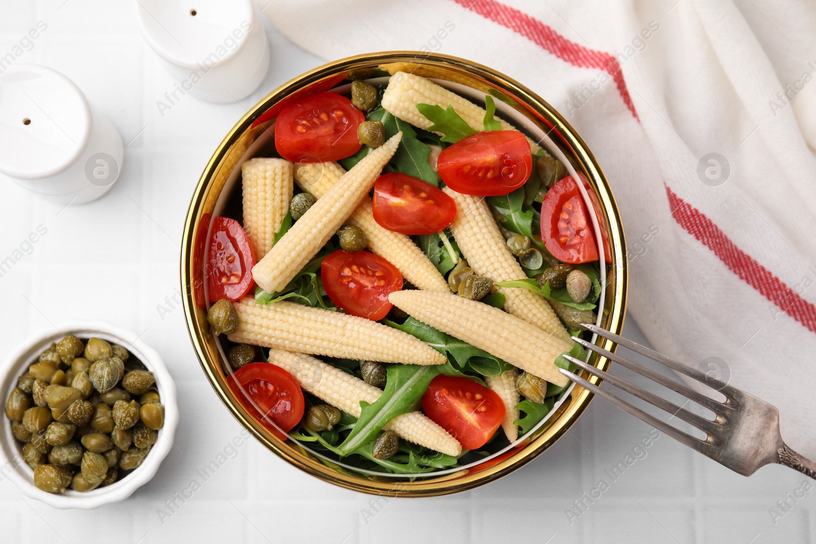 Photo of Tasty baby corn with tomatoes, arugula and capers on white tiled table, flat lay