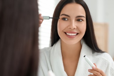 Photo of Beautiful woman applying serum onto her eyelashes indoors, reflection in mirror. Cosmetic product
