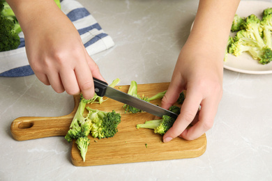 Photo of Woman cutting raw broccoli at light grey marble table, closeup