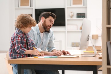 Photo of Man working remotely at home. Busy father talking on smartphone while his son drawing at desk