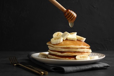 Photo of Pouring honey from dipper onto delicious pancakes with bananas and butter at dark table, space for text