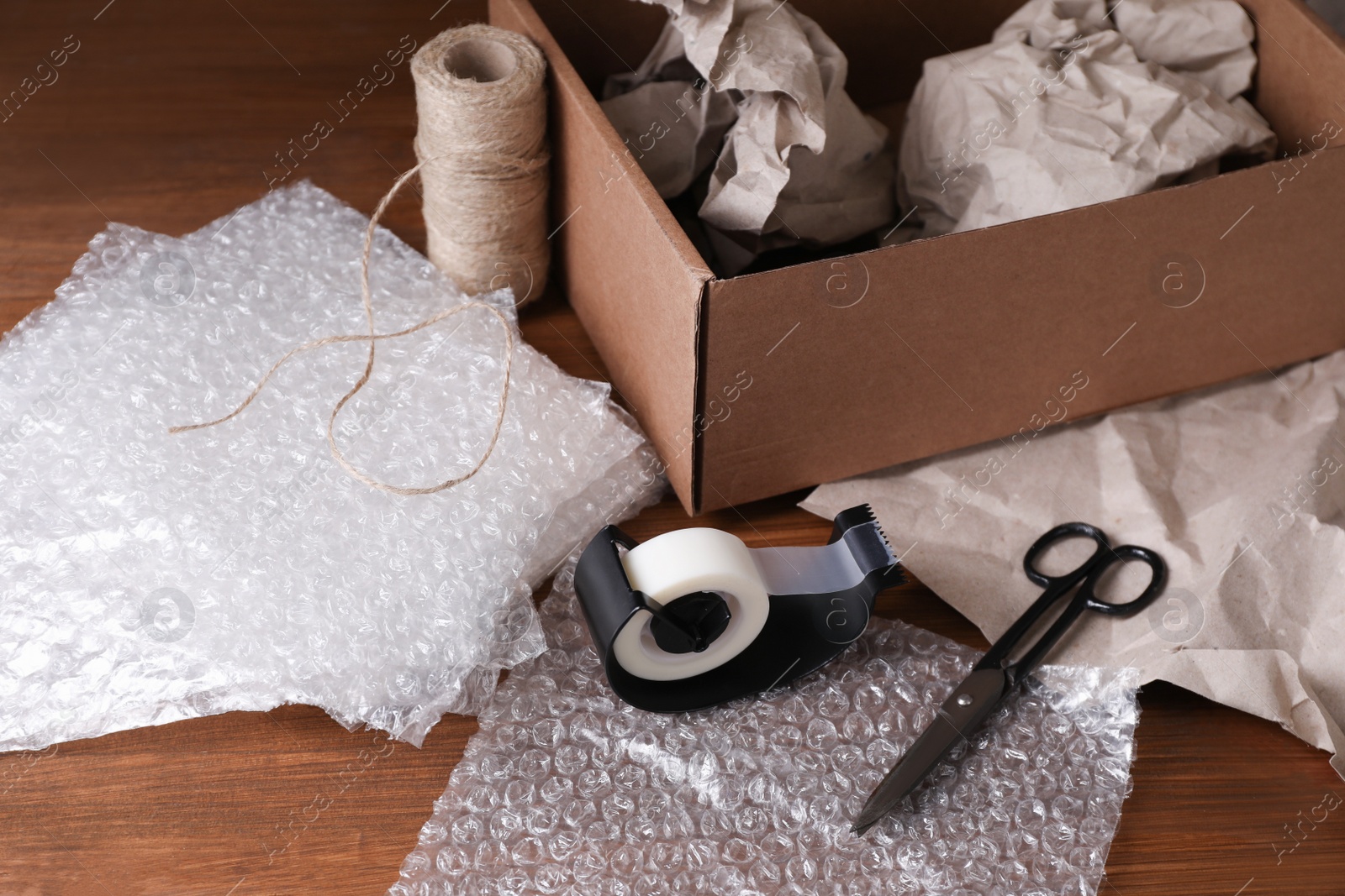 Photo of Bubble wrap, adhesive tape, twine and scissors near cardboard box on wooden table