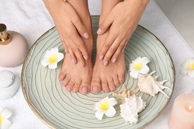 Photo of Woman soaking her feet in plate with water, flowers and seashells on white towel, closeup. Spa treatment