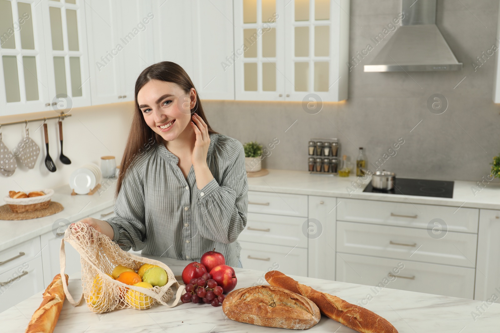 Photo of Woman with string bag of fresh fruits, baguettes and bread on light marble table