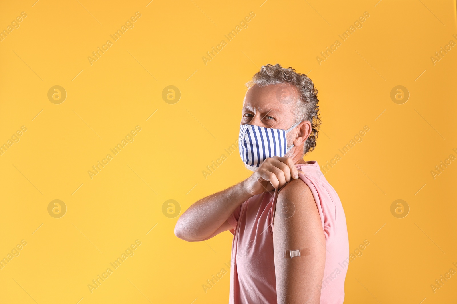 Photo of Senior man in protective mask showing arm with bandage after vaccination on yellow background. Space for text