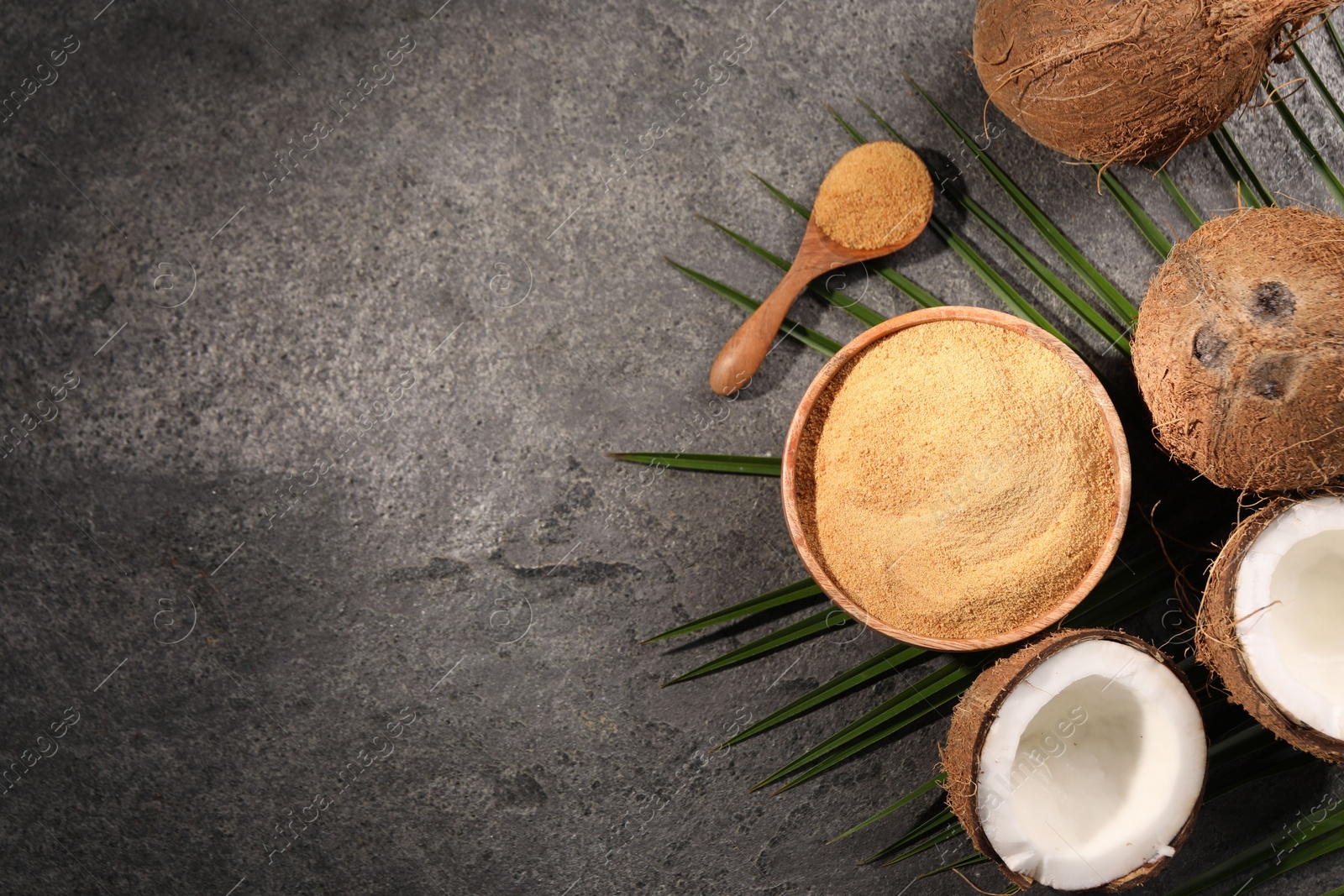 Photo of Coconut sugar in bowl, spoon, palm leaves and fruits on dark textured table, flat lay. Space for text