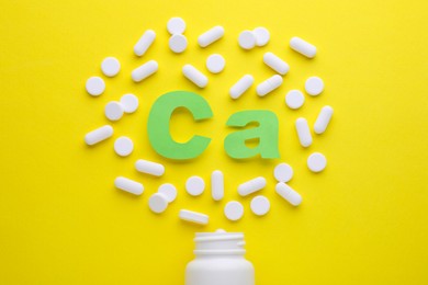 Photo of Pills, open bottle and calcium symbol made of green letters on yellow background, flat lay