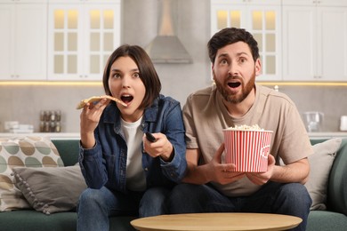 Photo of Surprised couple watching TV with popcorn and pizza on sofa at home