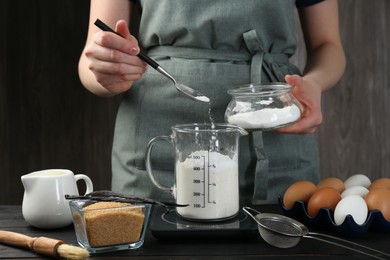 Woman adding baking powder into measuring cup at black wooden table, closeup