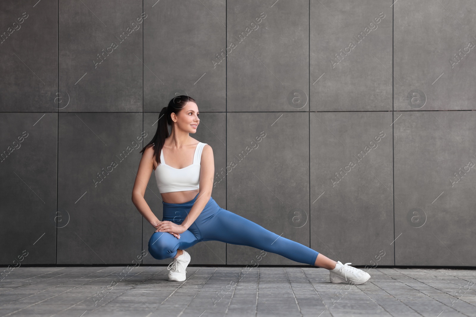 Photo of Smiling woman in sportswear stretching near dark grey wall outdoors