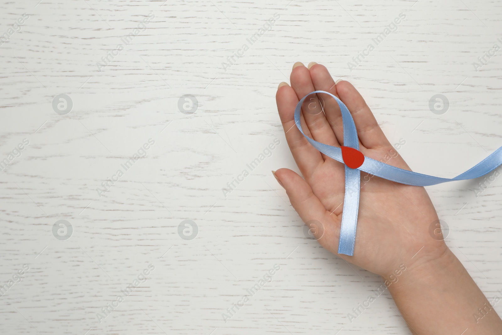 Photo of Woman holding light blue ribbon with paper blood drop at wooden table, top view and space for text. Diabetes awareness