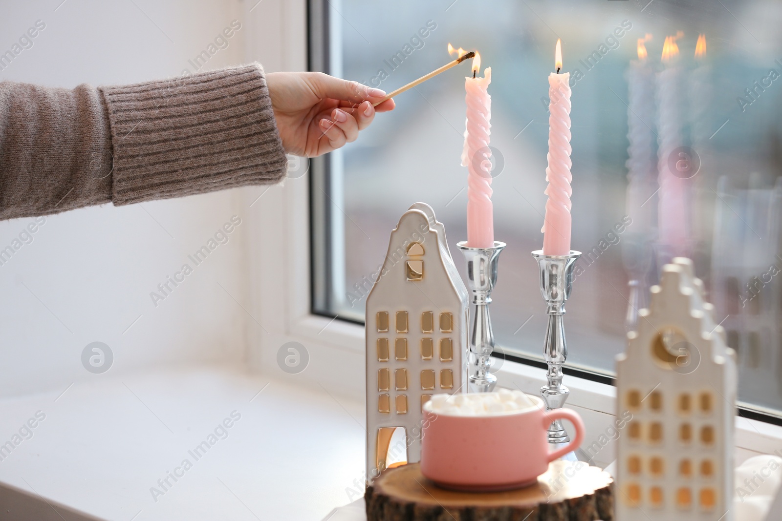 Photo of Woman lightning up candle near windowsill with house shaped lanterns indoors, closeup