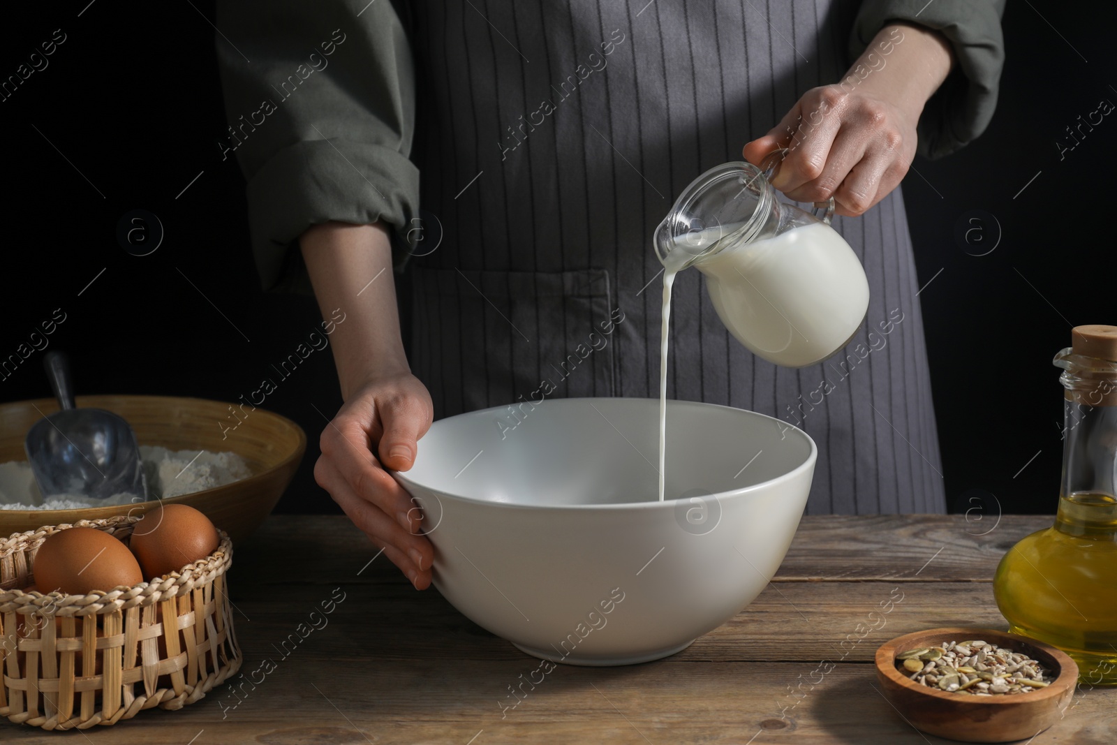 Photo of Making bread. Woman pouring milk into bowl at wooden table on dark background, closeup