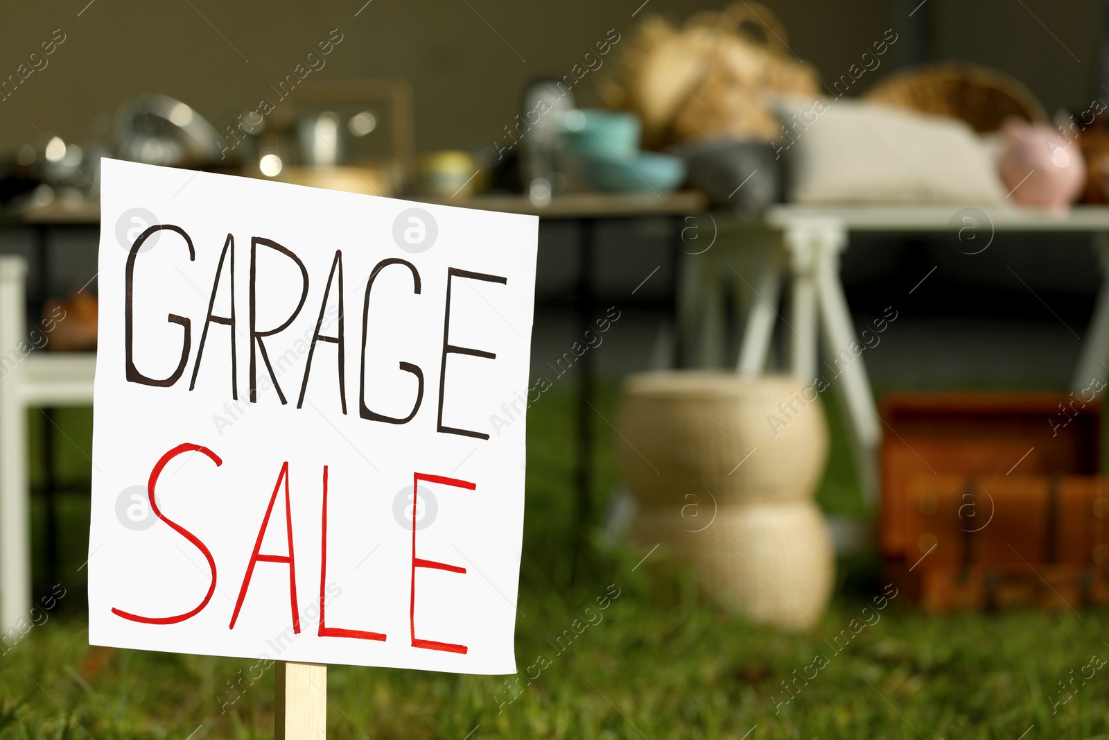 Photo of Sign Garage sale written on cardboard near tables with different stuff outdoors, closeup. Space for text