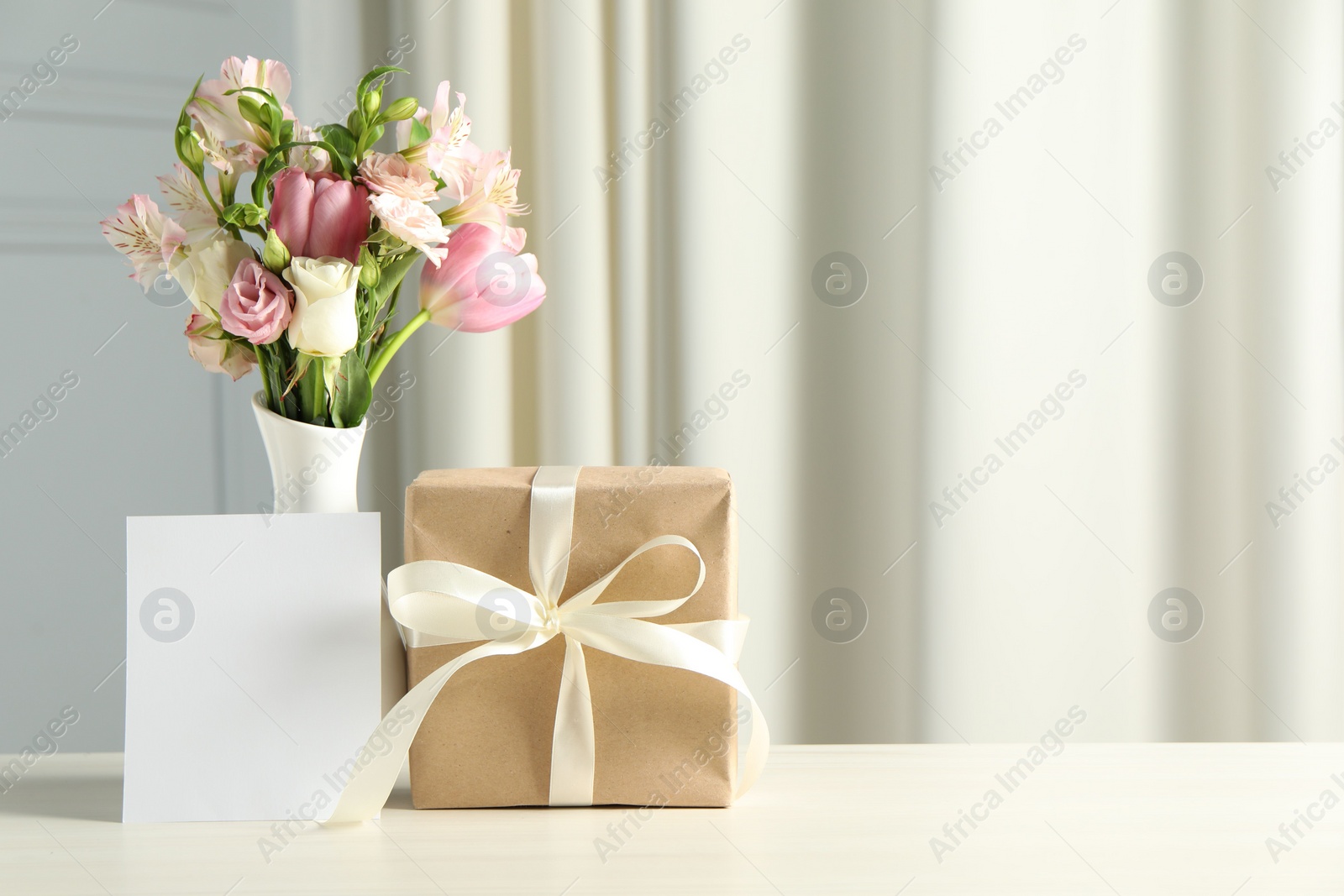 Photo of Happy Mother's Day. Gift box, blank card and bouquet of beautiful flowers in vase on white table indoors. Space for text