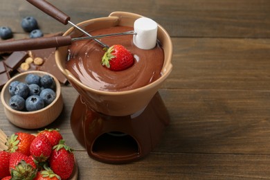 Photo of Fondue pot with melted chocolate, different fresh berries, kiwi, marshmallow and forks on wooden table, space for text