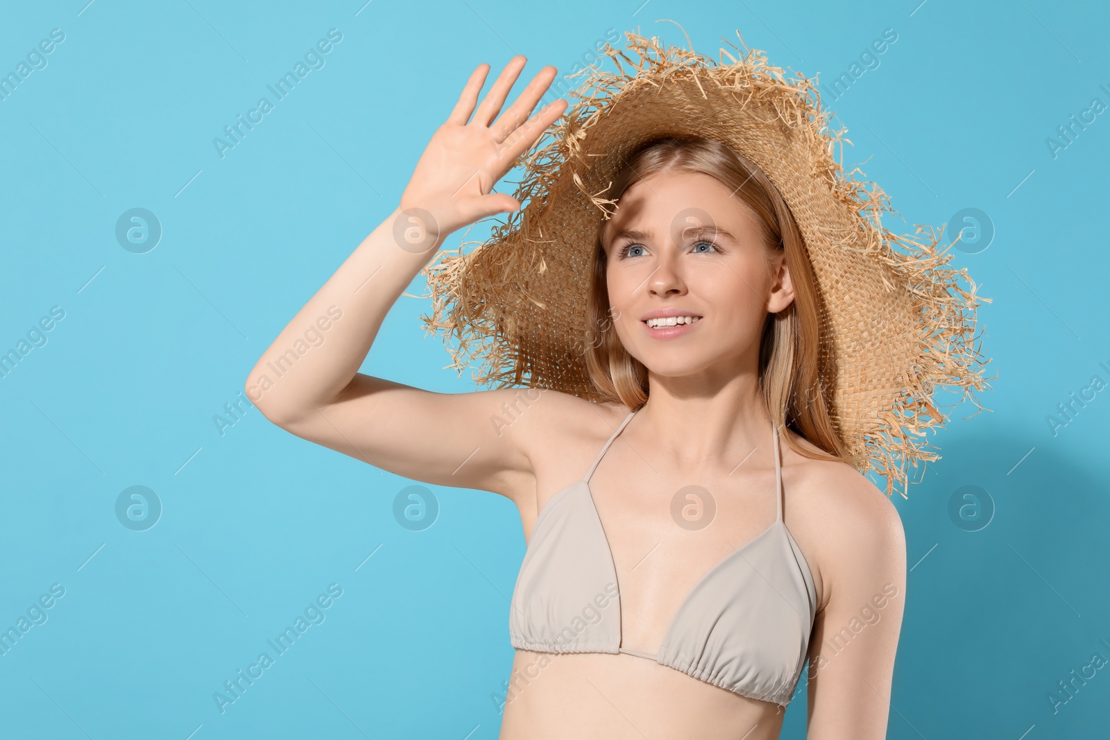 Photo of Beautiful young woman in straw hat shading herself with hand from sunlight on light blue background. Space for text