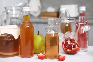 Tasty kombucha in glass bottles, jar and fresh fruits on white tiled table