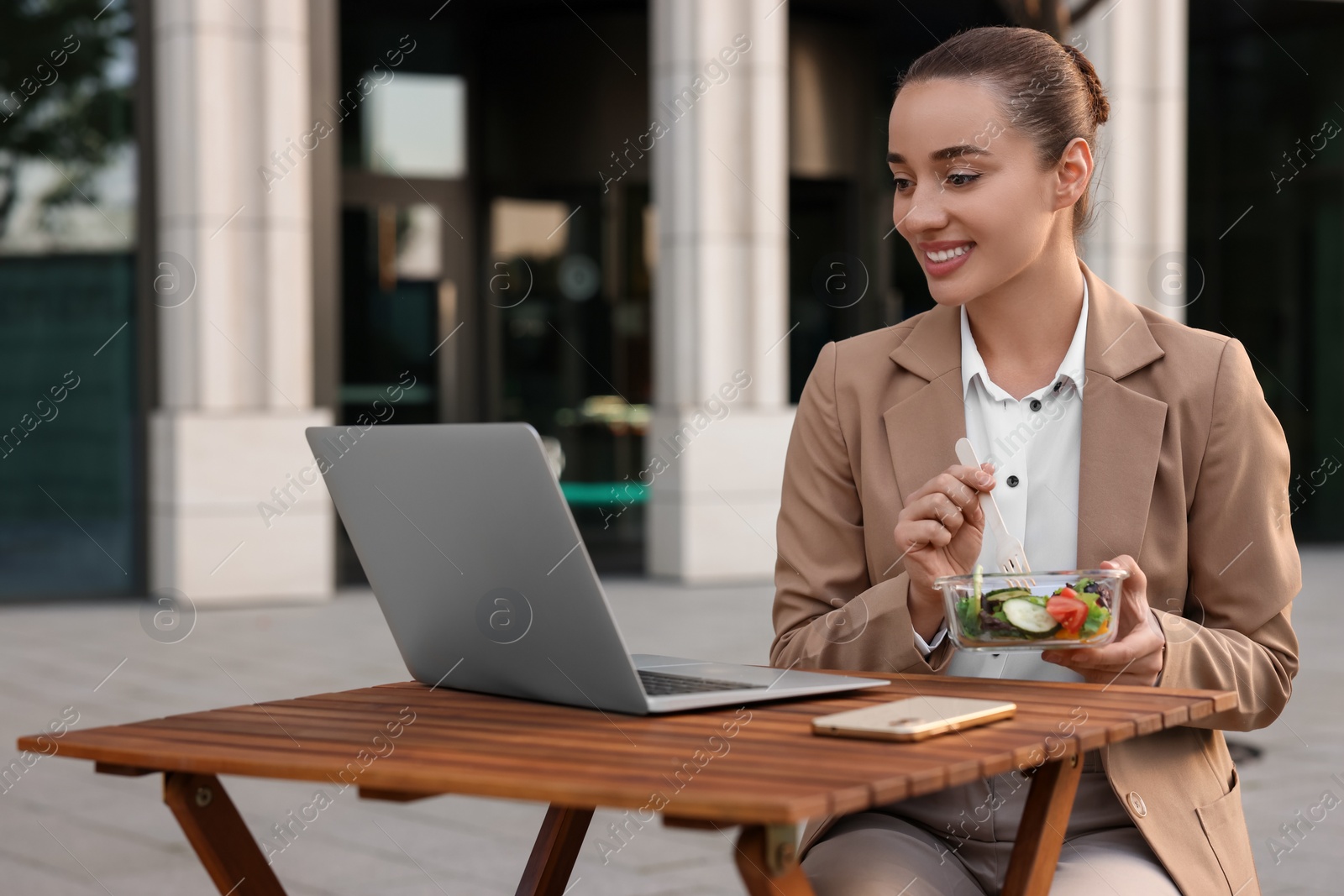 Photo of Happy businesswoman using laptop during lunch at wooden table outdoors