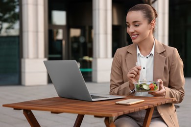 Photo of Happy businesswoman using laptop during lunch at wooden table outdoors