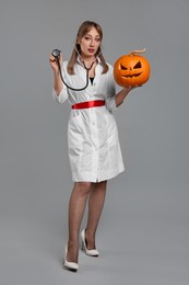 Photo of Woman in scary nurse costume with carved pumpkin and stethoscope on light grey background. Halloween celebration