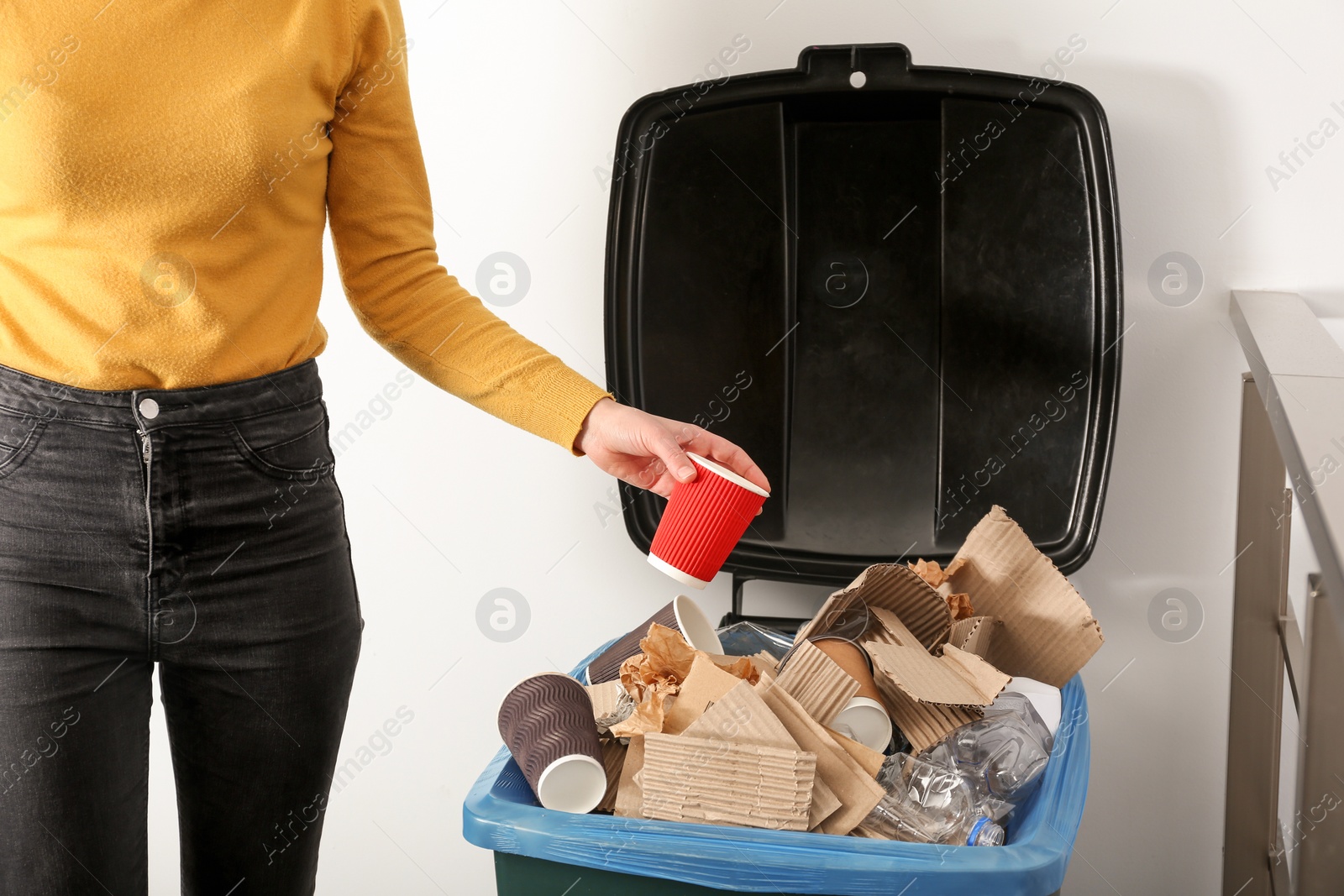 Photo of Young woman throwing coffee cup in trash bin indoors, closeup. Waste recycling