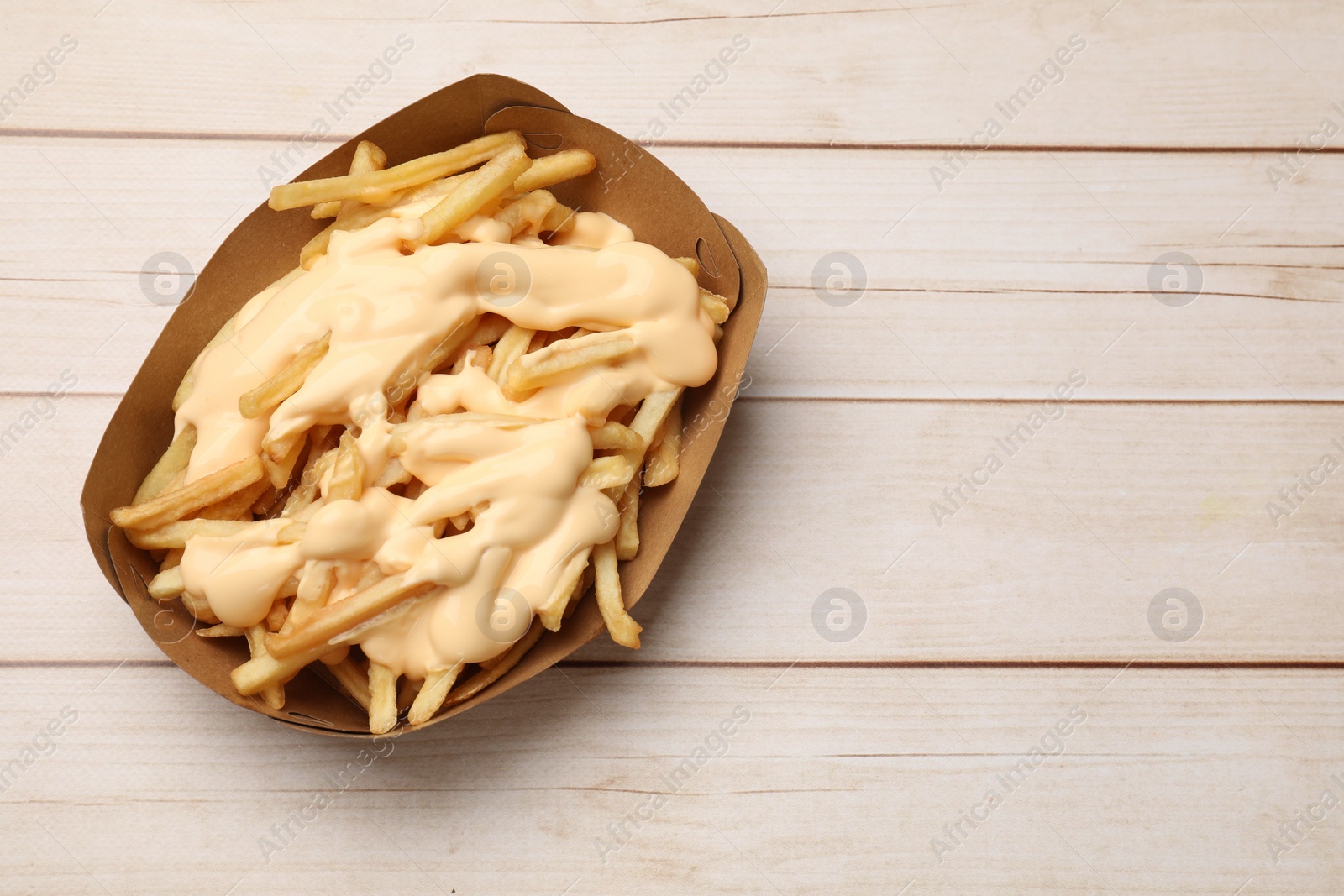 Photo of Tasty potato fries and cheese sauce in paper container on light wooden table, top view. Space for text