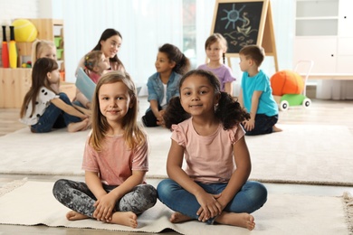 Photo of Cute girls sitting on floor while kindergarten teacher reading book to other children indoors