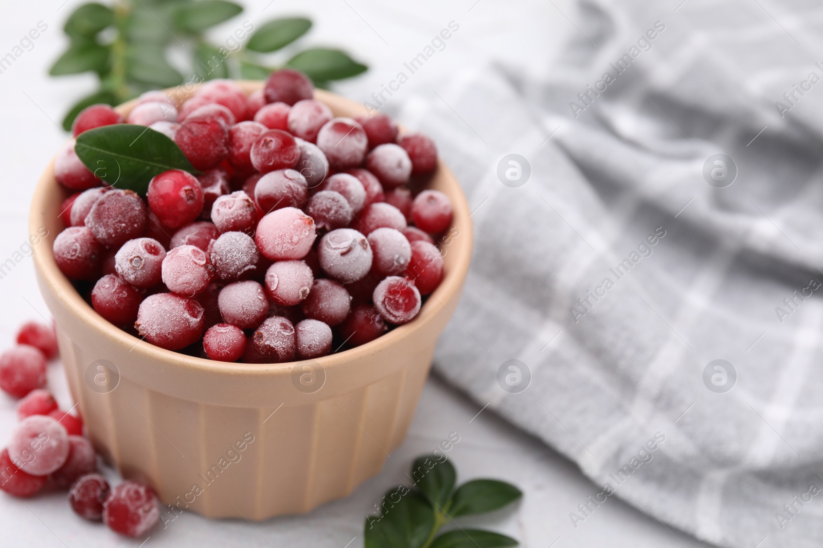 Photo of Frozen red cranberries in bowl and green leaves on white table, closeup. Space for text